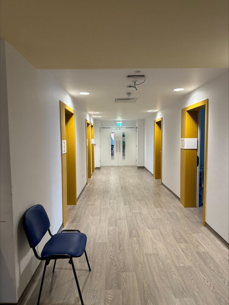 Aylesbury clinic 4 | NUPAS Long hallway with white walls, yellow-framed doors, and blue chairs at the left foreground; exit sign visible at the far end.